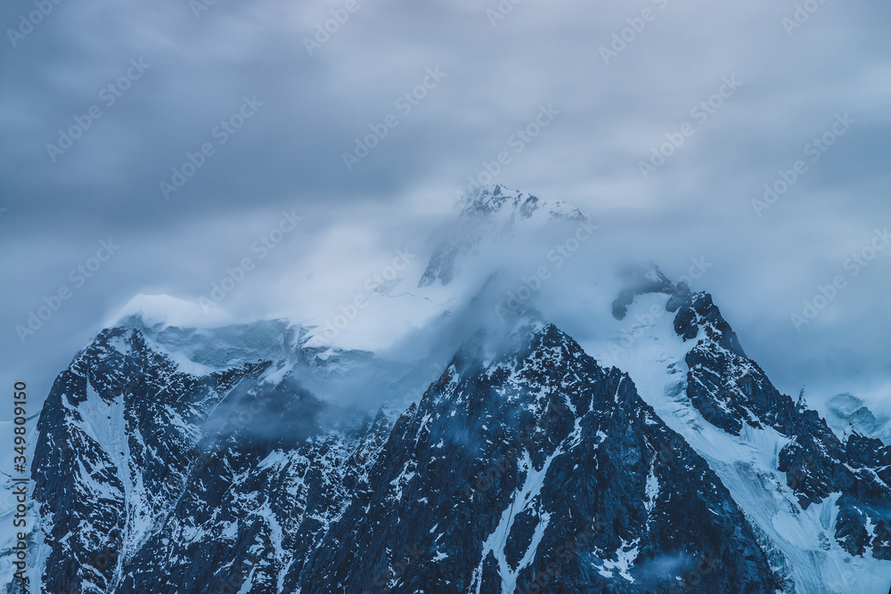 Mysterious dramatic alpine scenery with snowy mountain top inside low clouds in dusk. Bleak view to glacier in cloudy sky in twilight. Atmospheric minimalist landscape with snowy rocks in dense fog.