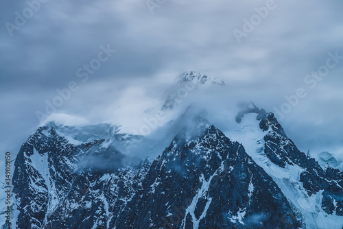 Mysterious dramatic alpine scenery with snowy mountain top inside low clouds in dusk. Bleak view to glacier in cloudy sky in twilight. Atmospheric minimalist landscape with snowy rocks in dense fog. © Daniil