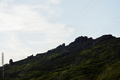 Silhouette of rocks in sunshine above cloudy sky. Vivid green mountain with crags in sunlight. Scenic minimalist alpine landscape in sunny day. Beautiful highland scenery with big stones on hillside.