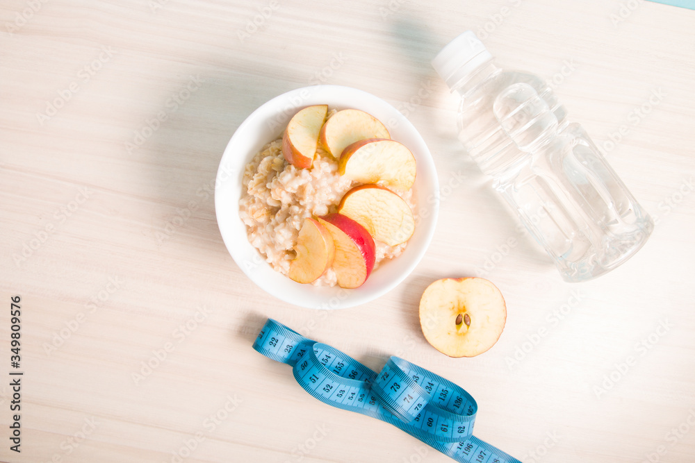 half apple, bottle of water, a bowl of boiled oatmeal with sliced apple and a blue measuring tape on a wooden table, proper nutrition concept, healthy breakfast half apple, bottle of water, a bowl of boiled oatmeal with sliced apple and a blue measuring tape on a wooden table, proper nutrition concept, healthy breakfast