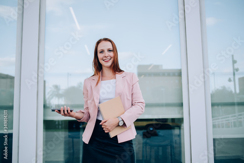 Free Download Prosperous redhead female manager holding copybook and mobile phone satisfied with successful career, portrait of smiling 20s business woman in elegant clothes looking at camera using gadget - GFXLoad