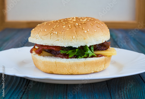 Burger of light bun with sesame, with greens, cheese, ketchup and meat cooked at home on white plate. Blue wooden background.