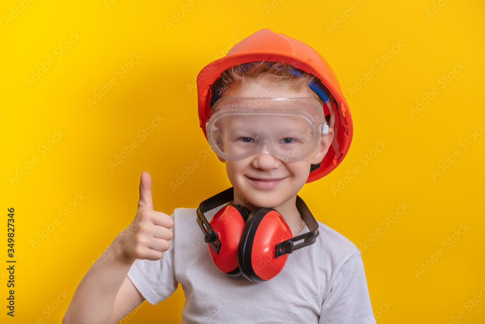 Cute boy in builder glasses, helmet and headphone to ear protection shown big finger up over yellow background copy space Cute boy in builder glasses, helmet and headphone to ear protection shown big finger up over yellow background copy space