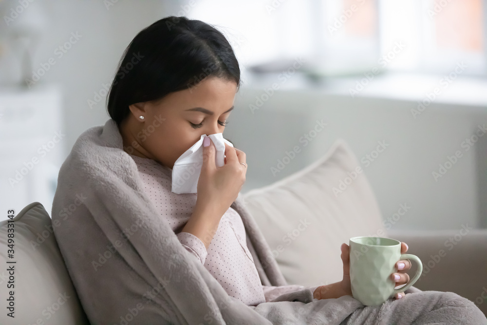 African woman sitting on sofa in living room wrapped in warm plaid hold paper handkerchief blow runny nose relief nasal stuffiness drinks cold medicine. Influenza flu symptoms, seasonal grippe concept African woman sitting on sofa in living room wrapped in warm plaid hold paper handkerchief blow runny nose relief nasal stuffiness drinks cold medicine. Influenza flu symptoms, seasonal grippe concept