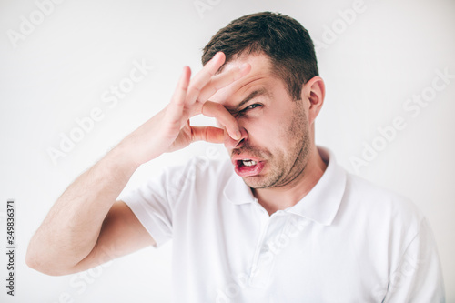 Young man isolated over white background. Guy covers his nose with fingers due to a bad smell. Look at camera with disgust.