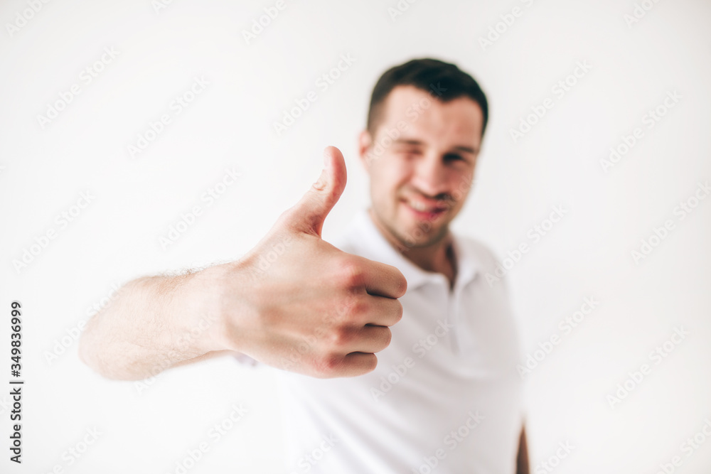 Young man isolated over white background. Guy hold one eye closed and show big thumb up. Like symbol. Cheerful attractive positive male person on blurred background. Young man isolated over white background. Guy hold one eye closed and show big thumb up. Like symbol. Cheerful attractive positive male person on blurred background.