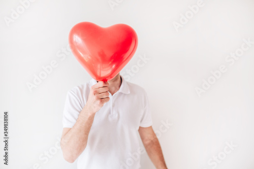Young man isolated over white background. Guy hold red balloon in heart shape in front face. Cover or hide it. Love.