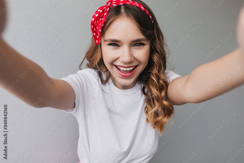 Sensual curly girl with red ribbon making selfie. Indoor photo of refined female model with wavy hairstyle laughing to camera. Sensual curly girl with red ribbon making selfie. Indoor photo of refined female model with wavy hairstyle laughing to camera.