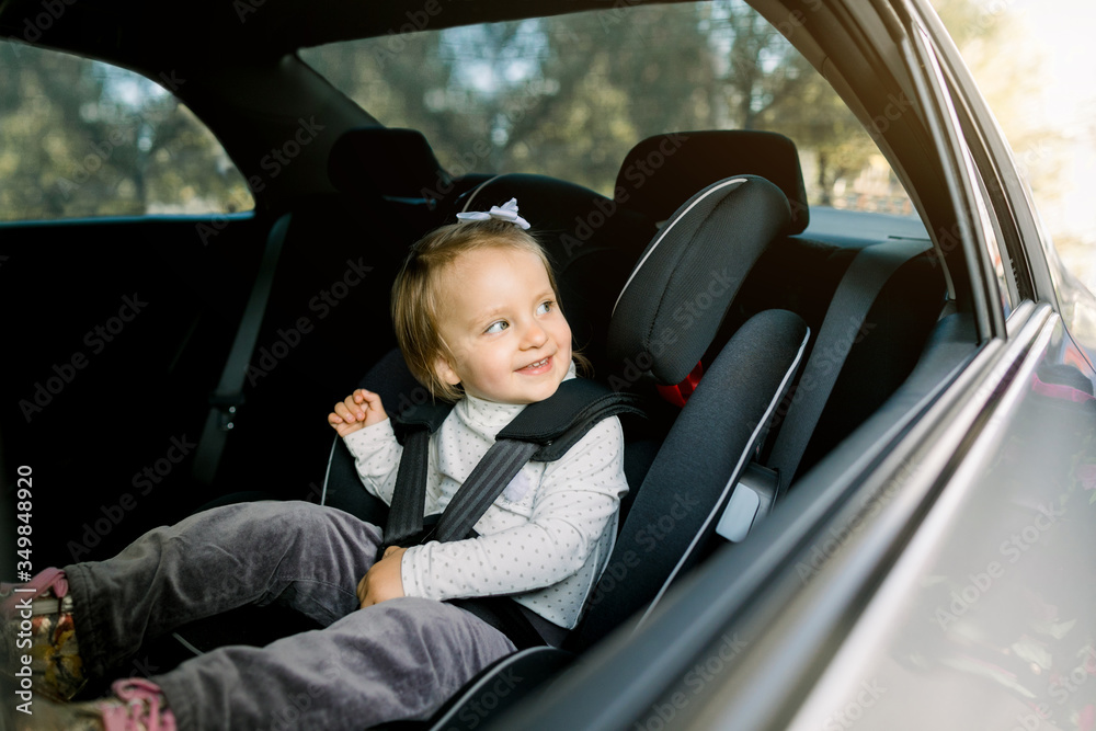 Beautiful smiling cute baby girl fastened with security belt in safety car seat, looking at the window. Child transportation safety concept