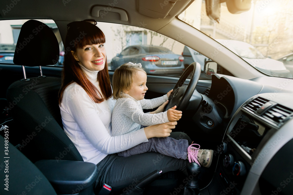 Young mother with her little baby girl sitting in the car in the front seat near car steering wheel. A woman and baby girl pretending to be driving. Mother shows her baby the car inside Young mother with her little baby girl sitting in the car in the front seat near car steering wheel. A woman and baby girl pretending to be driving. Mother shows her baby the car inside