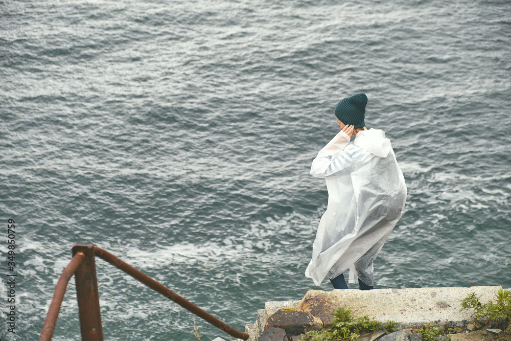 A young girl in a raincoat walks down an old rusty stairs on a steep cliff toward the sea on a foggy summer day. Lifestyle and outdoor concept.