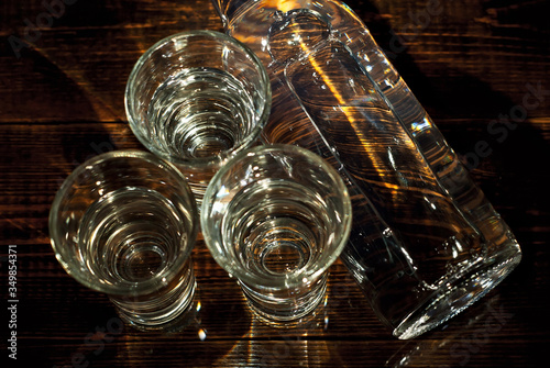 Glasses with alcohol near the bottle. Transparent drink on a brown wooden background. Table made of dark shabby boards. Blue reflexes and bright reflections of light on the bottle.