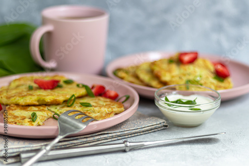 Zucchini fritters, vegetarian zucchini fritters, served with fresh herbs and sour cream. Garnished with strawberries and green onions. On a light gray background.