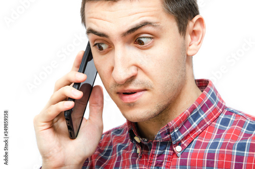 Young man in a plaid shirt with a smartphone on a white background. The guy holds a smartphone by the ear, talking on the phone. Anger, anger, surprise, attentiveness during a telephone conversation