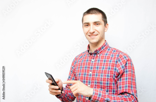 Young man in a plaid shirt with a smartphone on a white background. The guy looks at the camera and holds a smartphone in front of him and points, presses a finger into it