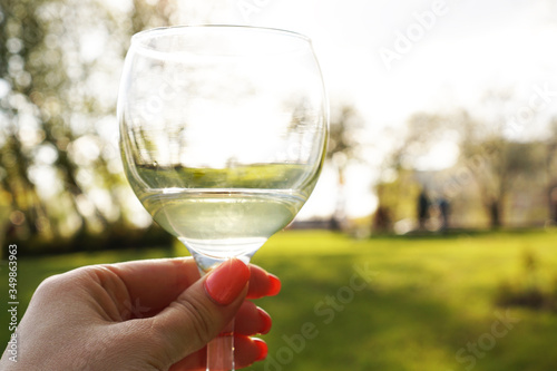 Woman hand holding a glass of white wine on the blurred background of green park at summer sunset