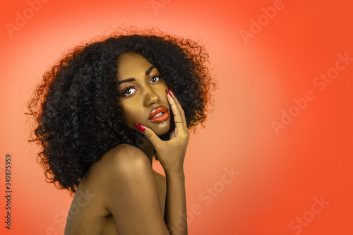 A sexy young black female with long black curly hair  beautiful makeup  popping orange lip stick   perfectly manicured nails posing by herself in front of an orange background.