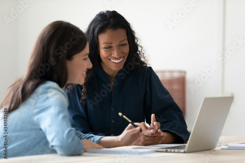 Close up happy young african american businesswoman using laptop with laughing female mentor in coworking boardroom at meeting. Smiling diverse woman managers talking about new business concept. photo