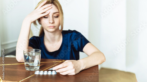 Cute woman holding her head and looking at a glass of water and pills. To feel bad. Coronavirus disease, covid-19. Feel sick