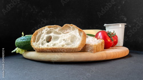 Two slices of white beautiful airy white bread, yogurt, fresh vegetables - cucumbers, tomatoes and celery, all on a round wooden cutting board on a black background close-up, side view.