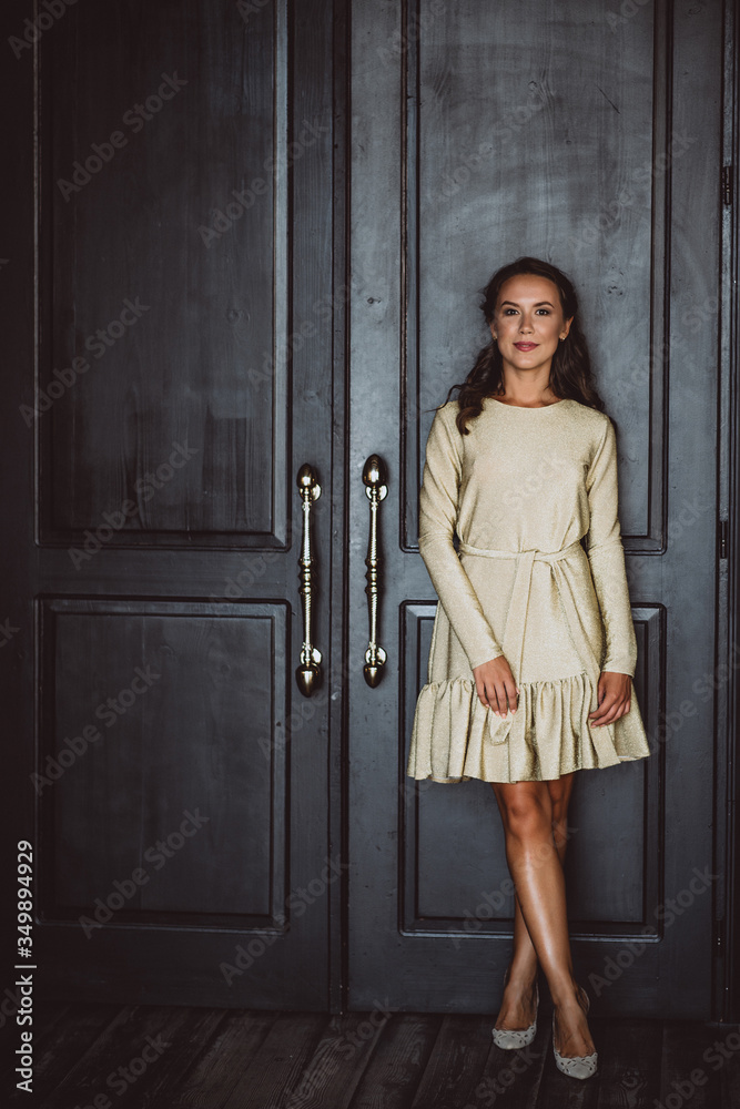 A beautiful young dark woman with dark curly hair and brown eyes in a gold dress is standing near the black door in the loft room. Soft selective focus.