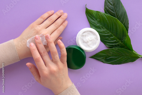 Young girls hand nearby white soft cream in jar with green leaves.Concept of eco cosmetic. Girl is moisturizing her hand by cream as beauty procedure at home.