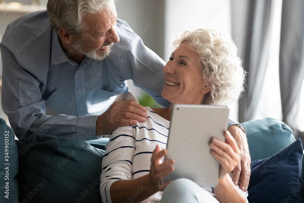 Happy grey-haired man embracing shoulders of pretty loving middle aged wife, communicating at home. Smiling mature woman using digital computer tablet, chatting talking with joyful husband indoors. Happy grey-haired man embracing shoulders of pretty loving middle aged wife, communicating at home. Smiling mature woman using digital computer tablet, chatting talking with joyful husband indoors.