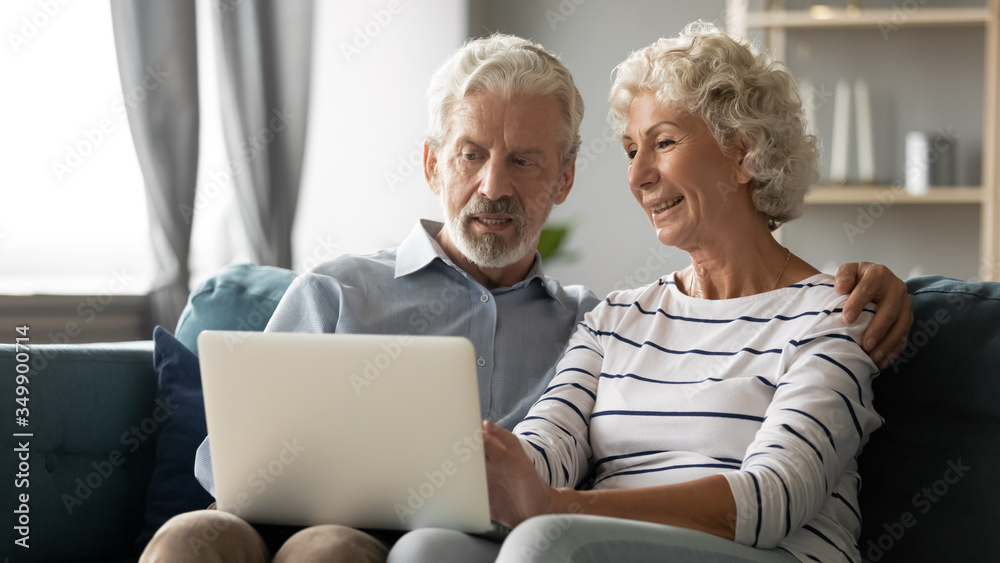 Happy middle aged family couple relaxing on sofa, using computer web surfing information shopping together at home. Elderly man showing laptop apps to retired wife, older generation with tech concept. Happy middle aged family couple relaxing on sofa, using computer web surfing information shopping together at home. Elderly man showing laptop apps to retired wife, older generation with tech concept.