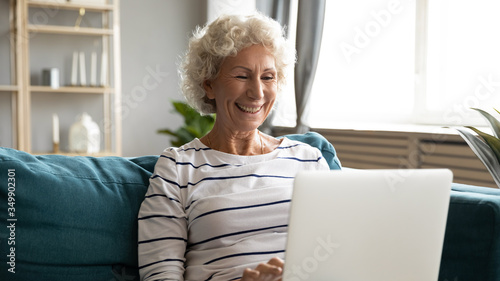 Happy hoary middle aged grandmother sitting on sofa, looking at laptop screen. Smiling grey haired mature woman reading pleasant news, chatting with friends in social network, watching funny movie.