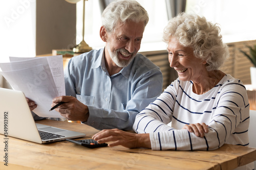 Happy middle aged family spouses sitting at table with computer and paper bills, calculating domestic incomes together at home. Smiling friendly mature senior married couple managing monthly budget.