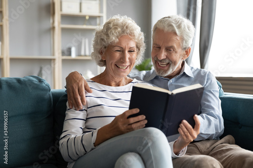 Happy middle aged man embracing attractive pleased mature hoary wife relaxing together on comfortable couch, enjoying reading favorite novel literature bestseller at home, retired people hobby.