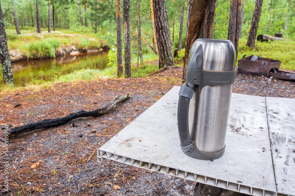 Beautiful camp thermos for hot tea stands on makeshift table in forest against background of taiga river. Beautiful camp thermos for hot tea stands on makeshift table in forest against background of taiga river.