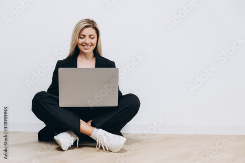 A beautiful young woman is sitting on the floor with a notebook pc on her lap, against a light background