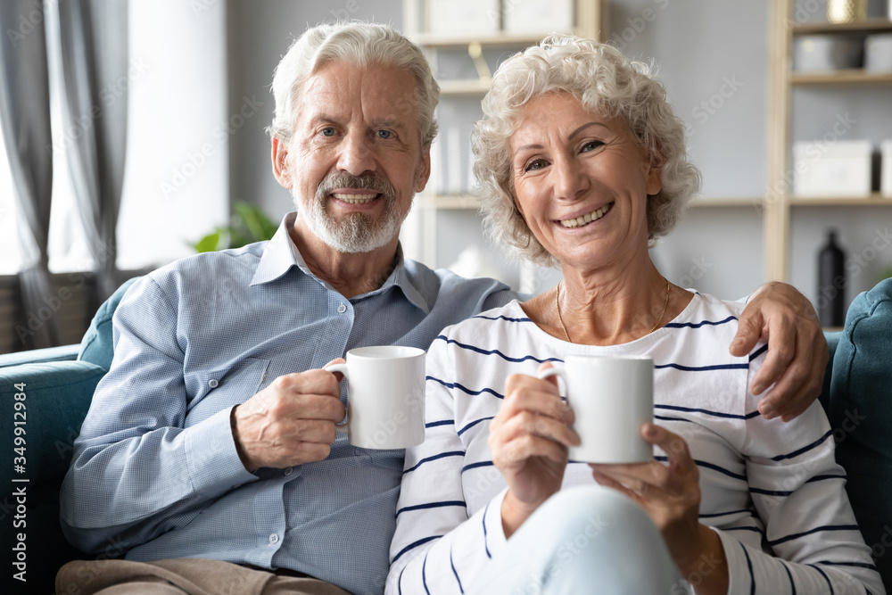 Portrait of happy middle aged married couple resting on comfortable sofa at home with cups of hot tea. Smiling retired family spouses enjoying lazy weekend morning time with coffee in living room. Portrait of happy middle aged married couple resting on comfortable sofa at home with cups of hot tea. Smiling retired family spouses enjoying lazy weekend morning time with coffee in living room.