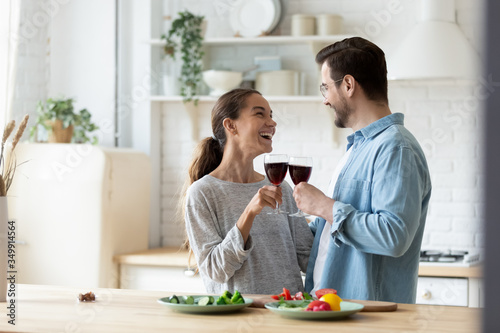 Overjoyed young husband and wife cling glasses drink wine celebrate anniversary or moving at home kitchen, happy millennial couple tenants cheers cooking preparing food together in modern new kitchen