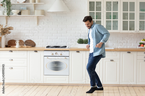 Happy millennial man enjoy leisure weekend dancing alone in bright modern design white kitchen, overjoyed young husband have fun entertain celebrate moving to new own home or apartment
