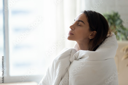 Calm millennial woman wrapped in blanket relax breathing fresh air at home, peaceful young female girl warm up in bed cover enjoy leisure weekend rest meditate indoors, stress free concept