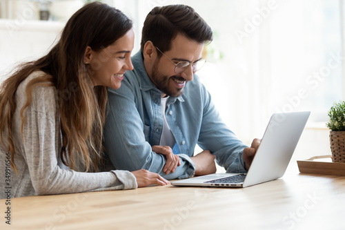 Happy millennial couple sit at table browsing wireless Internet on modern laptop together, smiling young husband and wife rest shopping online on computer, have fun with new technology device