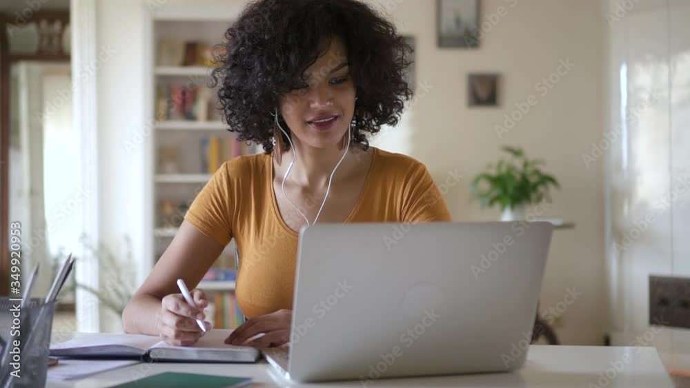 Young woman student using laptop to study online from home in internet Spbd. millennial girl writing in notebook looking at screen listen to teatcher and nod. headphones. distance learning quarantine