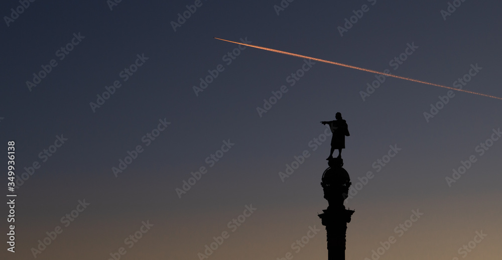 A plane crosses the sky over Barcelona at sunset as Columbus points in the right direction.