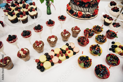 Table with lots of desserts, sweets, there are cake with cholocate and red and blue berries, macaroons, desserts with crram, fresh flowers photo