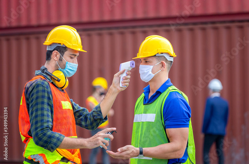 Factory worker man checking fever by digital thermometer for scan and protect from Coronavirus (COVID-19) at cargo containers - Healthcare Concept
