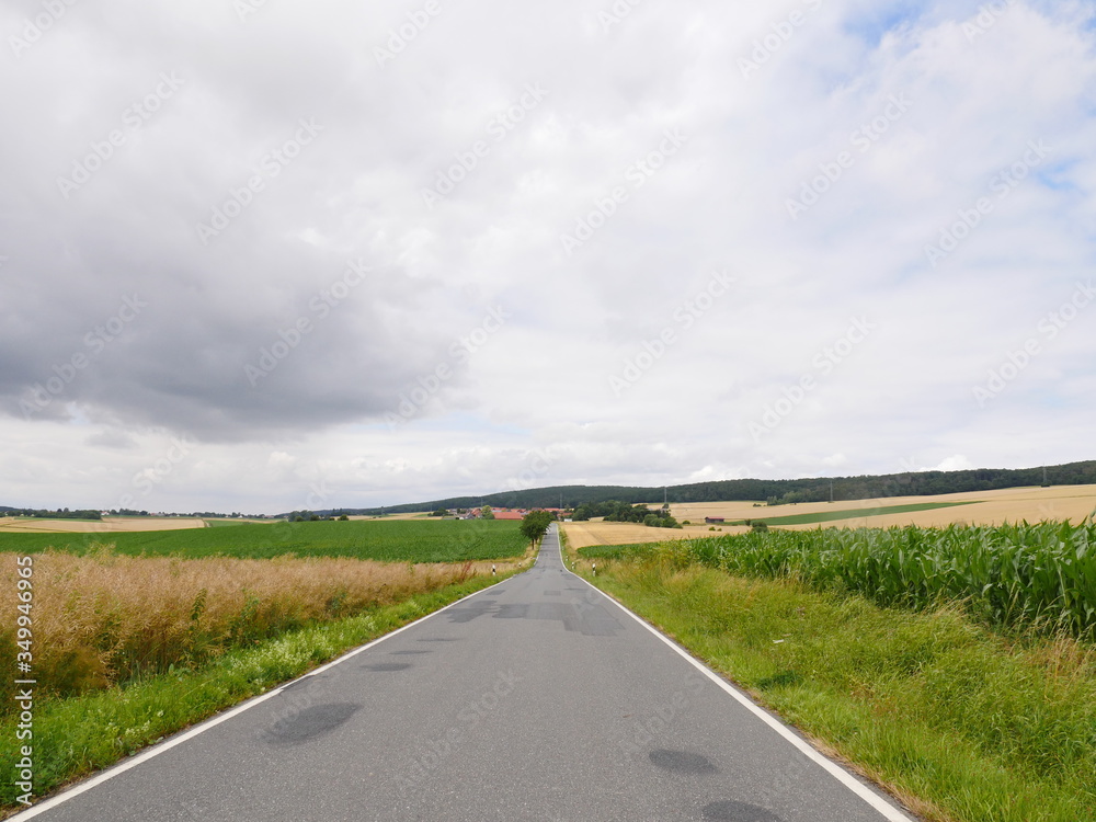 Asphalted repaired country road straight through hilly country towards the village of Helmscherode in the area of the hills from the Vorharz.  Fields of all kinds in the landscape