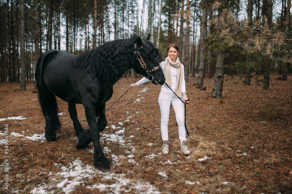 Magnetic young female in grey fur coat and white trousers stands with a big black horse outside Magnetic young female in grey fur coat and white trousers stands with a big black horse outside