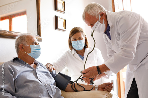 Anziano con mascherina viene visitato da un medico a casa che gli prende la pressione sanguigna insieme ad un altra dottoressa che assiste photo