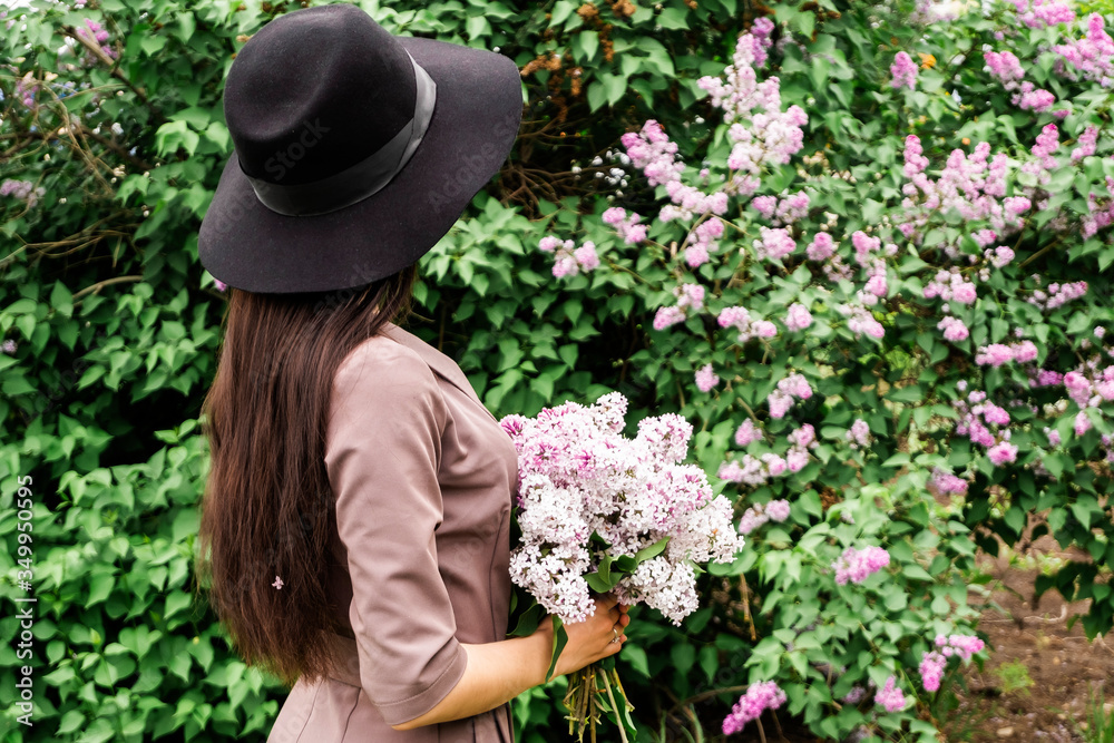Photoshoot in a lilac. A brunette in a black hat looks back at the lilac bush in a frame. Girl in a hat with a bouquet of lilac backs.
