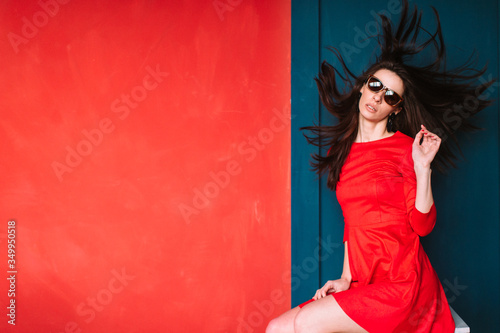 Beautiful fashion girl with long hair  spanish appearance in sunglasses and red elegnat dress posing on blue red wall in studio. Stylish brunette babe with flying hair. Professional hairstyle concept.