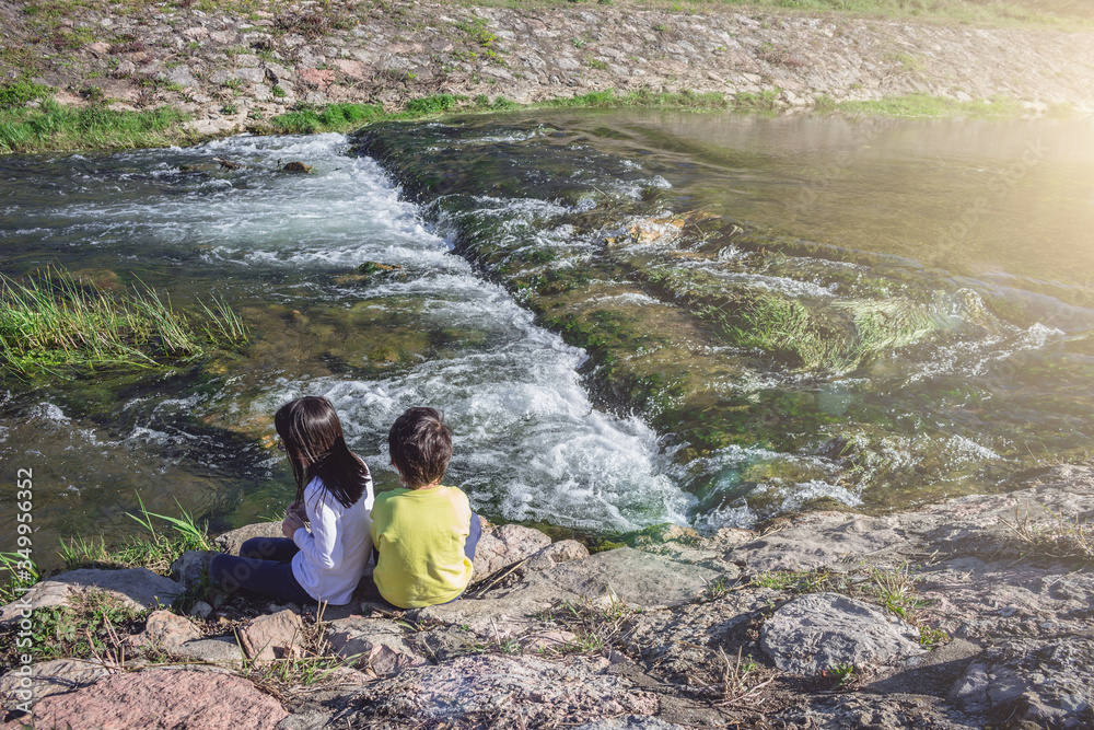 Children sitting by the river side. Two kids or siblings watching the fast water flow and playing outdoors. Stone embankment, exploring the world concept.