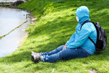 a man in a blue hooded jacket, jeans, a white mask and a gray backpack sits on the green grass on the river Bank in a Park .rest and freedom while walking. quarantine