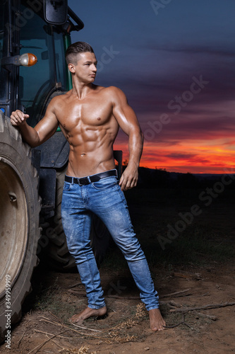 Muscular young sexy guy posing with a naked torso near the tractor in blue jeans at sunset outdoors. Muscular sexy young guy with athletic body posing at sunset.